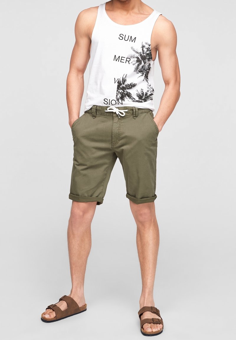 Débardeur blanc avec texte noir et graphique de palmier. Shorts vert olive avec ourlets retroussés. Sandales marron avec lanières et boucles.