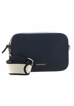 Petit sac bandoulière en cuir bleu marine avec une large bandoulière réglable en tissu noir et blanc et un logo COCCINELLE doré sur le devant.