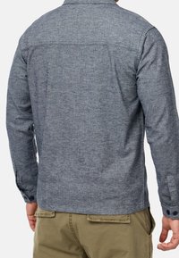 Homme portant une chemise à manches longues gris chiné et un pantalon kaki, vu de dos sur un fond uni.