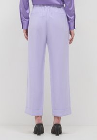 Persona che indossa pantaloni larghi lilla con orli risvoltati e una blusa di raso lilla abbinata, abbinata a scarpe con tacco nere, in piedi di fronte a una parete bianca.