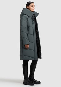 Padded long coat in dark green, featuring a tall collar and quilted design. Paired with black knee-high boots and slim-fit black leggings.