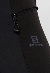 Černý sportovní top s kapsou na zip, hladkou texturou a výrazným šedým logem Salomon na boku. Materiál odvádějící vlhkost.