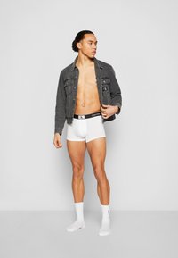 Baltos boksininkų šortai su juodai balta Calvin Klein juostele, derinami su pilku džinsiniu švarku. Modelis stovi neutralioje fone.
