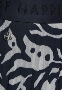 Černobílá vzorovaná látka s sklady, kovový kulatý knoflík a tmavý pás s vyvýšeným černým textem.