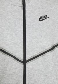 Sudadera gris con cremallera, hecha de una mezcla de algodón, con cremallera y acentos negros, y un pequeño logo negro en la parte superior izquierda.