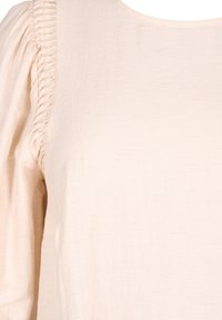 Blusa beige chiaro con una texture liscia, scollatura rotonda e dettagli arricciati sulle maniche. Tessuto morbido e leggero.