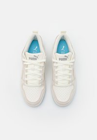 Para białych trampek Puma z beżowymi zamszowymi akcentami, białymi sznurowadłami i niebieskimi wkładkami z logo Puma, widziana z góry na białym tle.