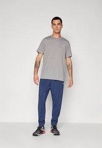 Mann med tatoveringer iført grå T-skjorte med korte ermer, marineblå joggebukser og svarte joggesko som står mot en ensfarget bakgrunn.