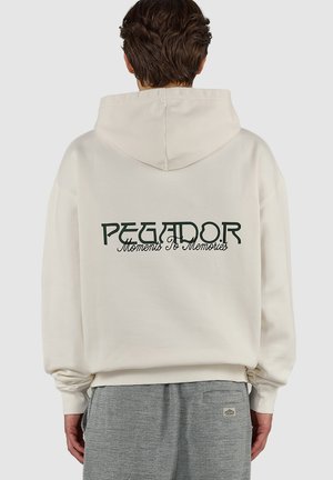 Personne portant un sweat à capuche crème avec le texte "PEGADOR Moments To Memories" dans le dos et un pantalon gris texturé, de dos à la caméra.
