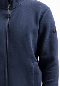 Donkerblauwe zip-up jas van textuurstof met een diagonale patroon, voorzien van een zijzak en een merkaccent in de vorm van een hexagoon op de mouw.