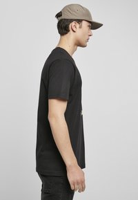 T-shirt en coton noir, à manches courtes, vue de profil latéral, associée à une casquette de baseball marron clair et un pantalon noir. Design simple et décontracté.