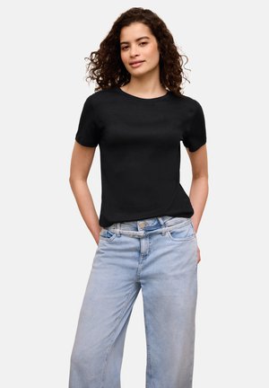 Nuori nainen, jolla on kiharat hiukset, pukeutuneena mustaan lyhythihaiseen T-paitaan ja vaaleansinisiin korkeavyötäröisiin farkkuihin, seisoo käsivarret taskuissa.