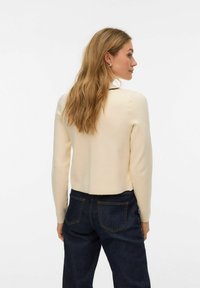 Kremowy sweter z długim rękawem o krótkim kroju. Gładka faktura, dopasowany w talii, zestawiony z ciemnymi dżinsami z tylnymi kieszeniami.