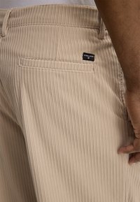 Zbliżenie na kieszeń beżowych spodni sztruksowych z metką Strellson na prawym biodrze, noszonych przez osobę o ciemnym odcieniu skóry.