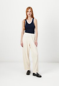 Tamsiai mėlynas tanktopas su baltu apvadu, derinamas su kreminėmis plačiomis kelnėmis. Juodi loafers'y baigia įvaizdį. Modelis laiko rudą petinę krepšį.