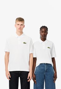 Białe koszulki polo z krótkim rękawem, z haftowanymi zielonymi logo krokodyla po lewej stronie klatki piersiowej, zestawione z ciemnymi dżinsami i szortami dżinsowymi.