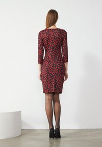 Figurbetontes Kleid mit Dreiviertelärmeln, rote Punkten auf schwarzem Hintergrund, knielang, glattes Material, kombiniert mit schwarzen High Heels.