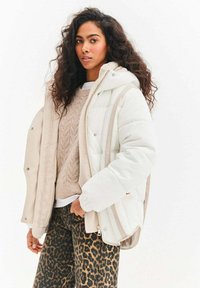 Kurtka puchowa w kolorze białym i beżowym z zamkiem błyskawicznym, wyposażona w kaptur i prążkowatą fakturę. Nosi się ją na beżowym, dzianym swetrze oraz spodniach w panterkę.