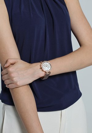 Donna che indossa un orologio da polso in oro rosa con quadrante bianco e cronografo al polso sinistro, vestita con un top senza maniche blu navy e pantaloni bianchi.