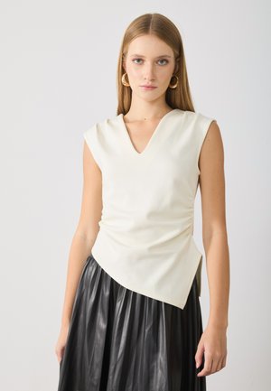 Young woman with straight blonde hair wearing gold hoop earrings, sleeveless white V-neck top, and black pleated skirt against plain background.