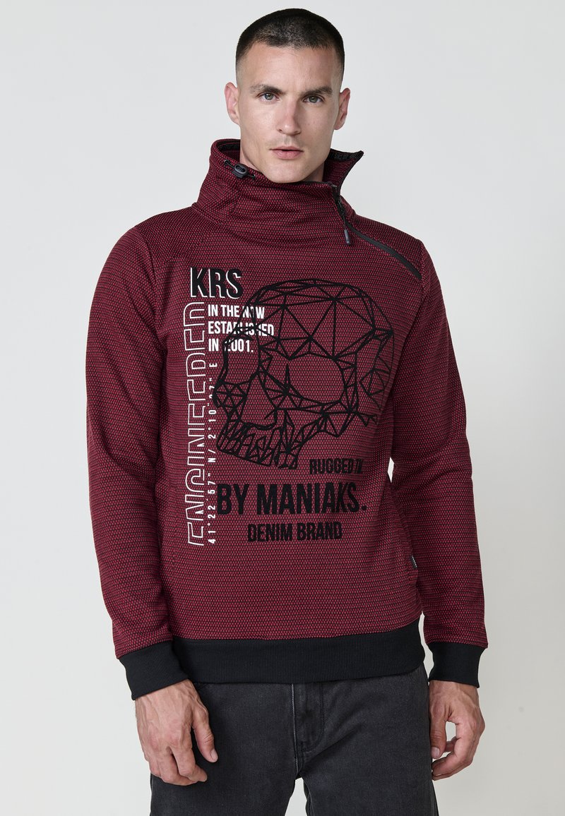 Sudadera roja texturizada con cuello alto, hardware negro y estampado gráfico que presenta un cráneo geométrico y tipografía en negrita en la parte delantera.