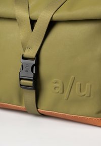 Olivgrünes Gewebe-Tasche mit strukturierter Oberfläche, schwarzer Kunststoffschnalle und braunem Lederbesatz. Mit geprägtem "a/u"-Logo.