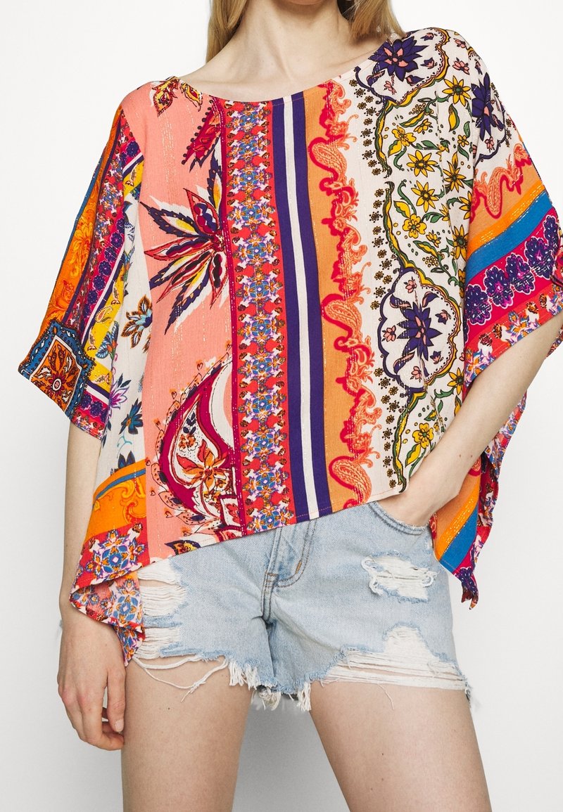 Femme portant un haut ample coloré à fleurs et rayures avec un short en denim clair déchiré, une main dans la poche, sur fond blanc.