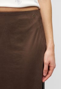 Καφέ σουέντ φούστα με ομαλή υφή, που διαθέτει μια μικρή χρυσή λεπτομέρεια λογότυπου "G" στη μέση και απλό, λιτό σχέδιο.