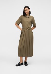 Μίντι φόρεμα χακί με κοντά μανίκια, μπροστινό άνοιγμα με κουμπιά, πλισέ φούστα και ζώνη στη μέση. Φορεμένο με μαύρα φλατ παπούτσια.