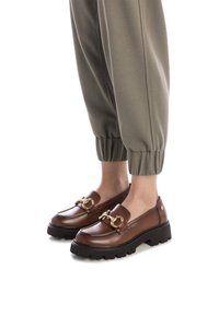 Mocasines de cuero marrón con una suela negra gruesa, con un detalle de cadena dorada en la parte frontal. Combinados con pantalones entallados en un tono oliva claro.