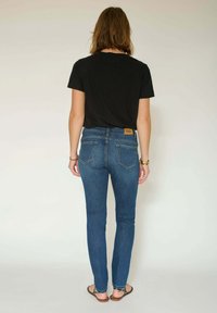 T-shirt noir, jean skinny bleu avec poches arrière et tongs. Le jean présente une légère décoloration et une étiquette sur la ceinture arrière.