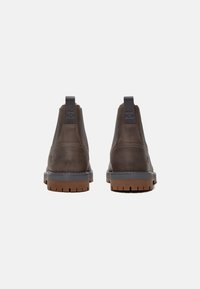 Hnědé kožené kotníkové boty s elastickými bočními panely, gumovou podrážkou a texturovaným povrchem. Zadní pohled ukazuje poutko pro snadné obouvání.