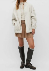 Lichtgrijze oversized jas met zilveren drukknoopjes, beige ribgebreide top, tan plissé rok en zwarte laarzen tot de knieën met een glanzende afwerking.