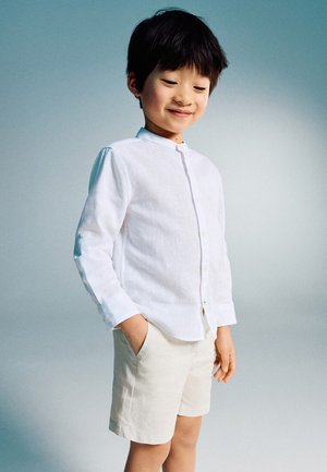 Young boy wearing a white long-sleeve buttoned shirt and light-colored shorts, standing with one hand in pocket, smiling slightly, against a plain background.