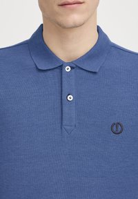 Blå polo skjorte laget av teksturert stoff med krage, som har to hvite knapper og en liten brodert logo på venstre side.