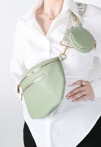 Marsupio in pelle verde a granulato con due scomparti con zip. La cintura decorativa regolabile presenta un motivo geometrico e hardware dorato.