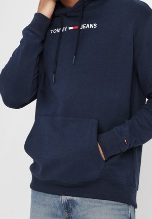 Person mit marineblauem Tommy Jeans Hoodie mit Fronttasche und hellblauer Jeans, eine Hand in der Tasche, die andere Hand in Kinnnähe.