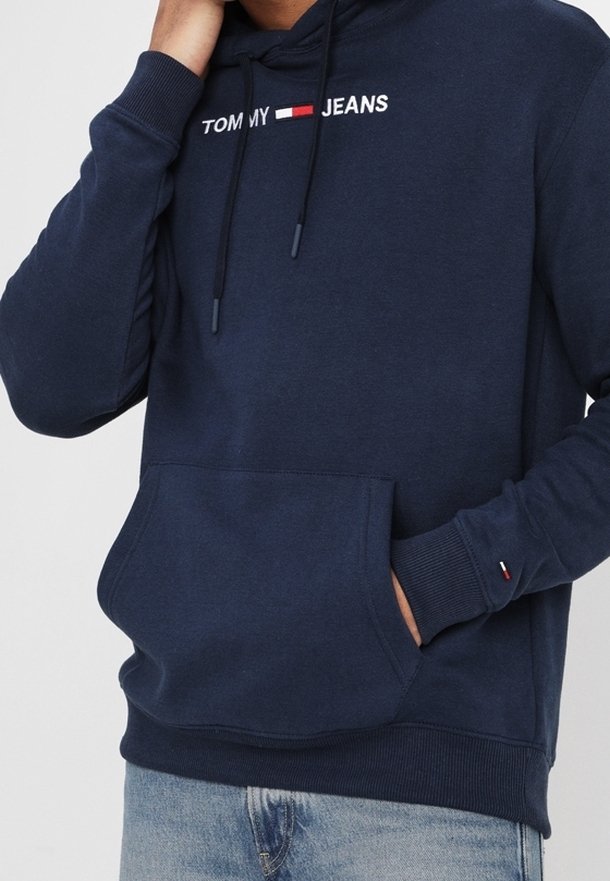Personne portant un hoodie Tommy Jeans bleu marine avec une poche frontale et un jean bleu clair, une main dans la poche, l'autre près du menton.