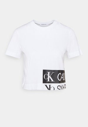 Biały krótki t-shirt z krótkim rękawem, krótki, z czarnym prostokątnym logo z napisem „cK CALVIN KLEIN JEANS” blisko dolnego brzegu.
