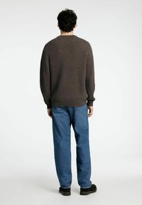 Bruine geribbelde trui met ronde hals, gecombineerd met lichtblauwe recht gesneden jeans. Het model staat met de handen langs het lichaam, gezien van achteren.