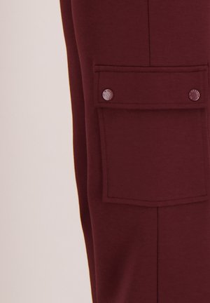 Close-up van bordeaux stoffen broek met een rechthoekige klepzak, vastgemaakt met twee ronde metalen drukknopen.