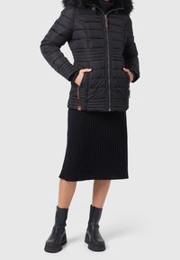 Chaqueta acolchada negra con capucha de pelo sintético, cremallera frontal y bolsillos laterales. Lleva puesta una falda negra acanalada hasta la rodilla y botines negros gruesos.