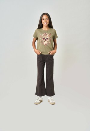 Fille aux longs cheveux bruns portant un t-shirt vert olive avec un motif de crâne fleuri, un pantalon évasé foncé et des baskets blanches, debout les mains dans les poches.