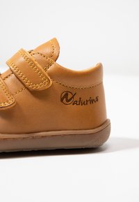 Hnědé kožené boty na suchý zip, s zaoblenou špičkou a gumovou podrážkou; mají logo vyražené na boku a kontrastní žluté prošívání.