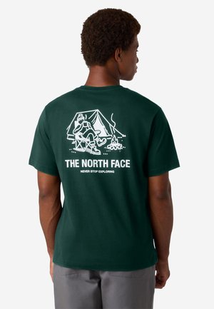Tummanvihreä puuvillapaita, jossa on valkoinen grafiikka leirintäautosta, teltasta ja nuotiosta selkäpuolella. Logoteksti "THE NORTH FACE" on alla.