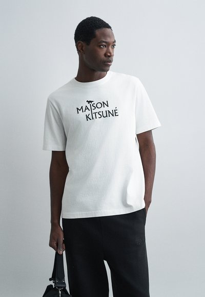 T-shirt bianco in cotone con colletto tondo, caratterizzato da un logo nero stampato "MAISON KITSUNÉ" sul davanti. Maniche corte, vestibilità comoda.
