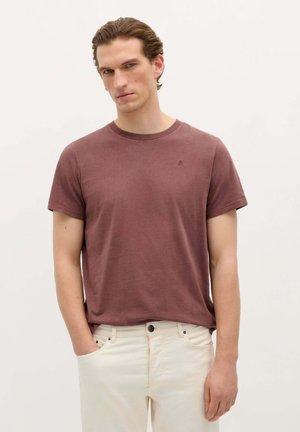Camiseta corta de manga en color burdeos, hecha de una tela suave, con un cuello redondo y un pequeño logo en el pecho, combinada con pantalones de color claro.