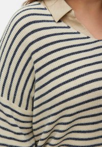 Gros plan d'un pull en maille beige avec de fines rayures horizontales bleu marine et un col beige.