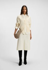 Cream cardigan over a white shirt, paired with a textured white skirt and black knee-high boots. A brown handbag completes the outfit.