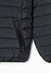 Sección de chaqueta acolchada negra con textura acolchada, que cuenta con un bolsillo lateral con cremallera y un acabado de tela suave.
